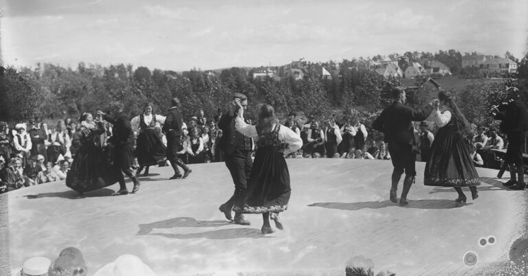 Mange samles for å danse sammen. Alle er i ført bunad. De danser under åpen himmel, langstrakt landskap.