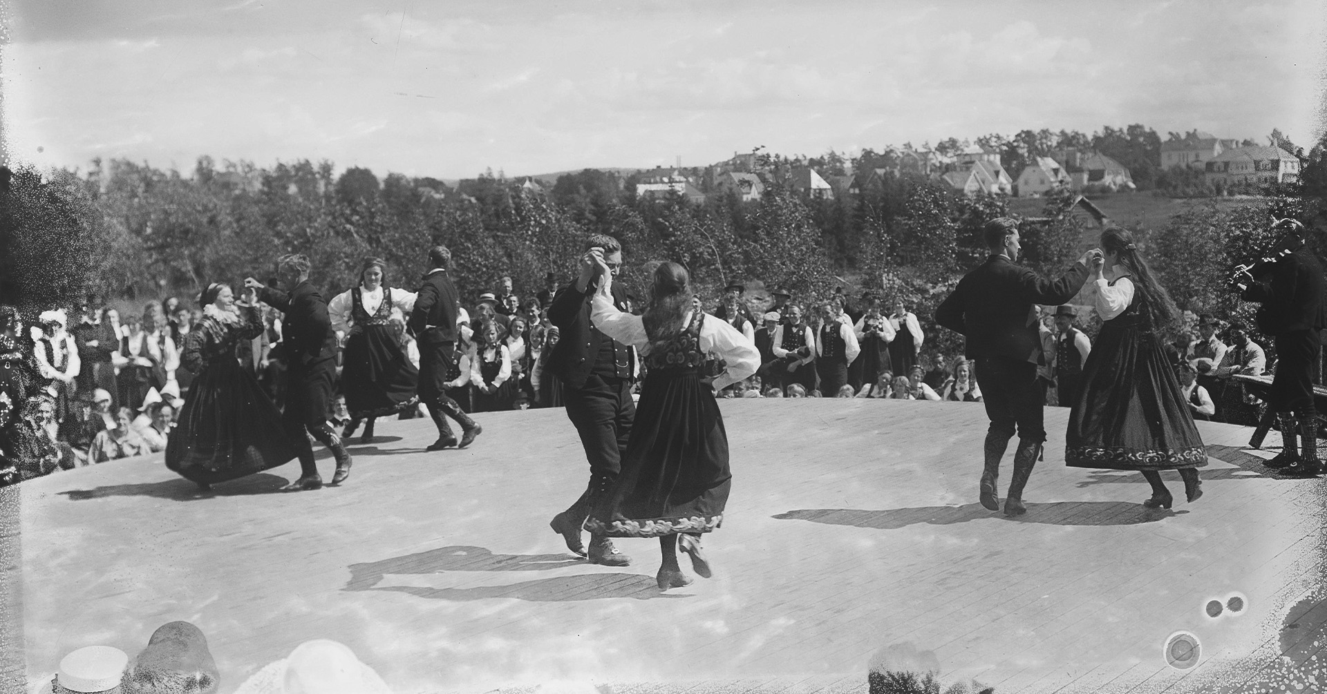 Mange samles for å danse sammen. Alle er i ført bunad. De danser under åpen himmel, langstrakt landskap.